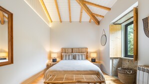 A bedroom with a large bed, wooden headboard, bedside lamps, a window with a view, and a woven basket. - Villa 243 - Charming Villa in a peaceful area (Orbacém)