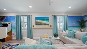 A living room with a large TV, a sofa, a coffee table, and a beach-themed mural on the wall.