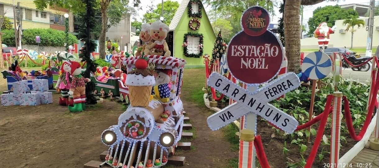 A festive outdoor scene with a decorated train, a sign reading "ESTAÇÃO NOEL," and a Christmas tree.