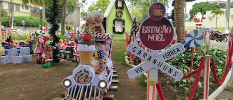 A festive outdoor scene with a decorated train, a sign reading "ESTAÇÃO NOEL," and a Christmas tree.