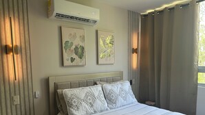 A bedroom with a bed, air conditioning unit, wall-mounted artwork, curtains, and a bedside table with a plant. - Charming 2-bedroom apartment in delightful Barranquilla with AC (Barranquilla)