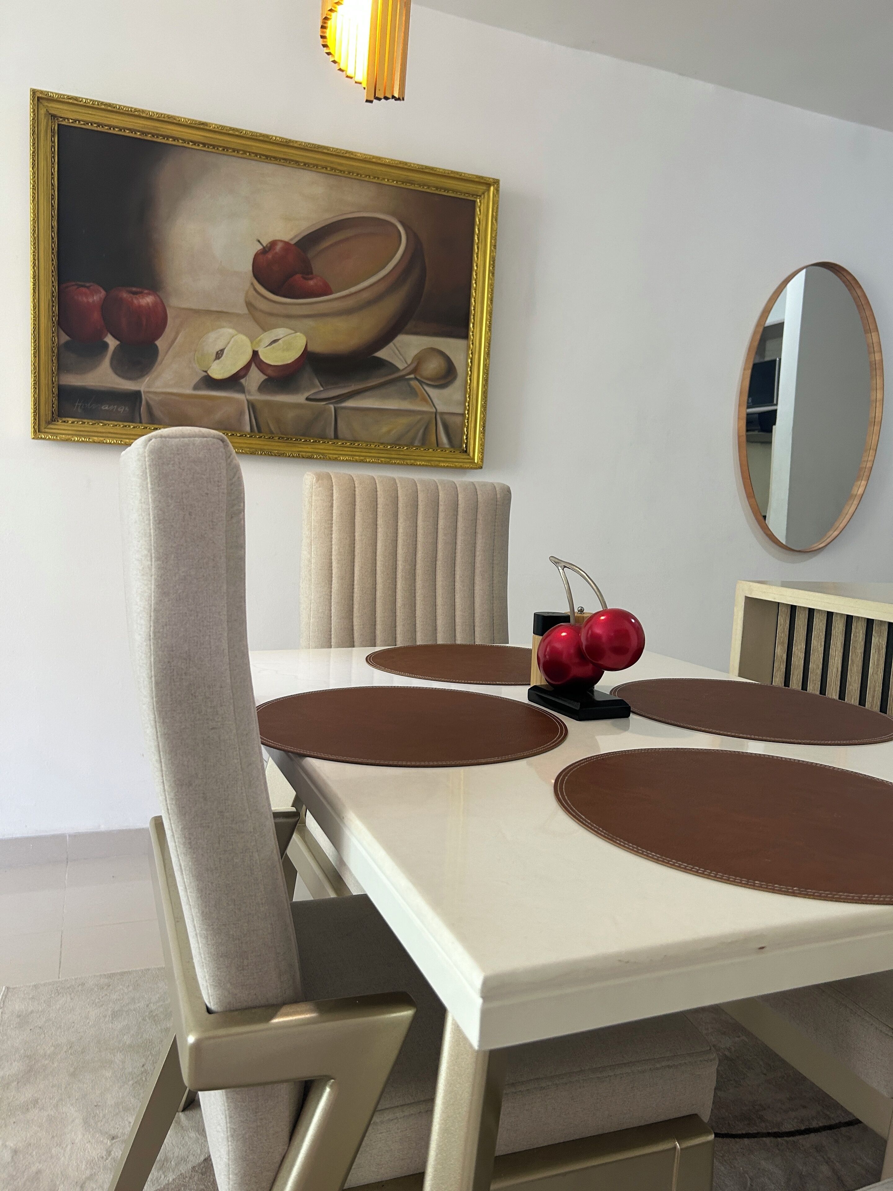 Un coin repas avec une table blanche, des sets de table marron et une pomme rouge posée sur un présentoir noir. Un tableau encadré est accroché au mur et un miroir se trouve au-dessus de la table.