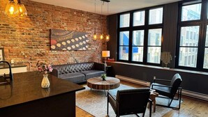 A modern living room with a brick wall, a sofa, a round coffee table, and a large window with a view of buildings. - Le Moyne-Heritage  (Montréal)