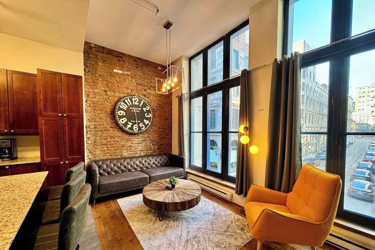 Un salon moderne avec un mur en briques, une grande horloge, un canapé, une table basse et une vue sur la ville grâce à de grandes fenêtres.