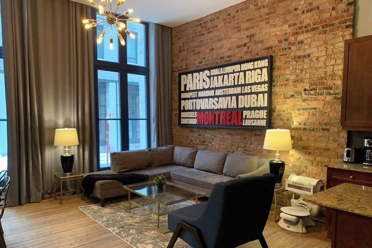 Un salon moderne avec un mur en briques, une grande fenêtre, un canapé d’angle, une table basse et un coin cuisine équipé d’un four à micro-ondes.