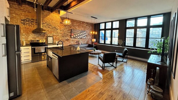 A modern kitchen with a large island, a brick wall, and a view of the city through large windows. - Le Moyne-Heritage  (Montréal)