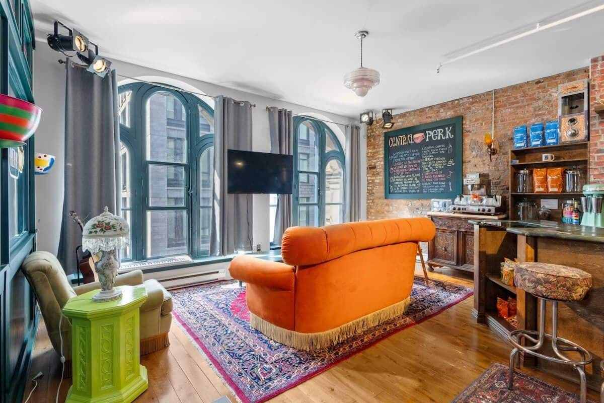 Un salon confortable avec une grande fenêtre, un téléviseur, une petite table d’appoint verte et un coin bar avec des tabourets.