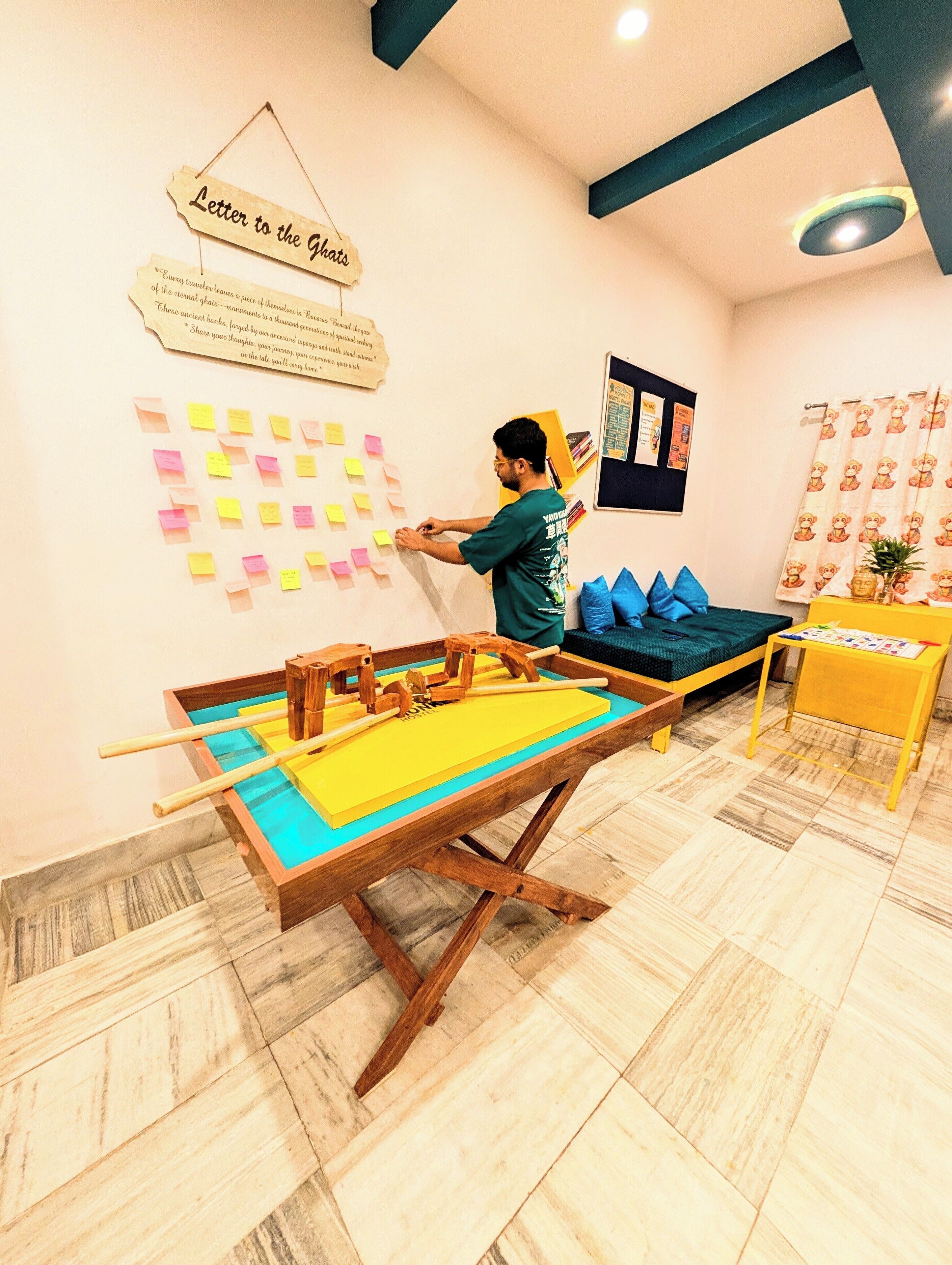A room with a pool table, a wall with sticky notes, a bed with blue pillows, and a small table with a plant.
