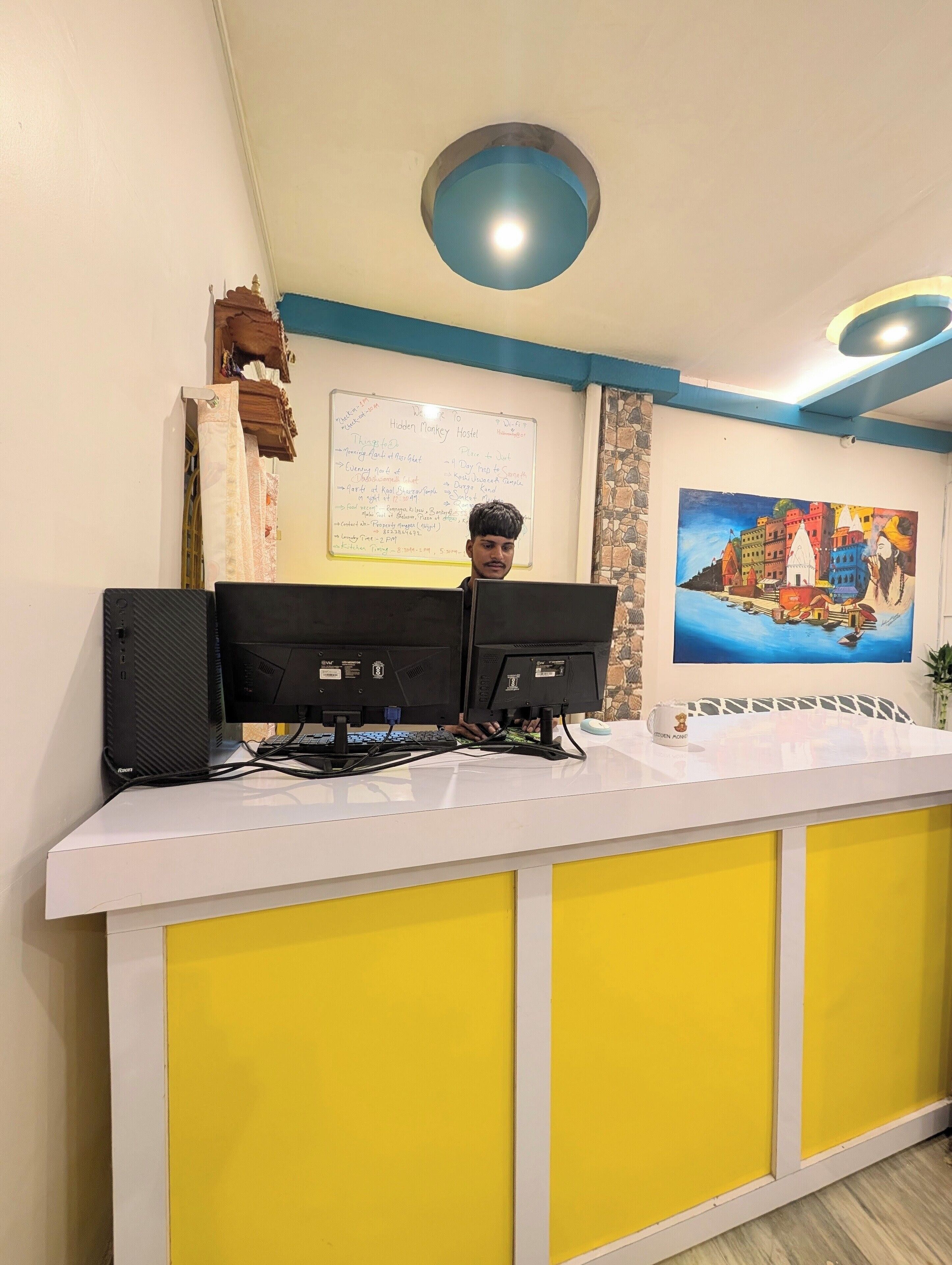 Un comptoir d’accueil équipé de deux écrans, un tableau blanc affichant des informations et un tableau représentant un paysage côtier.