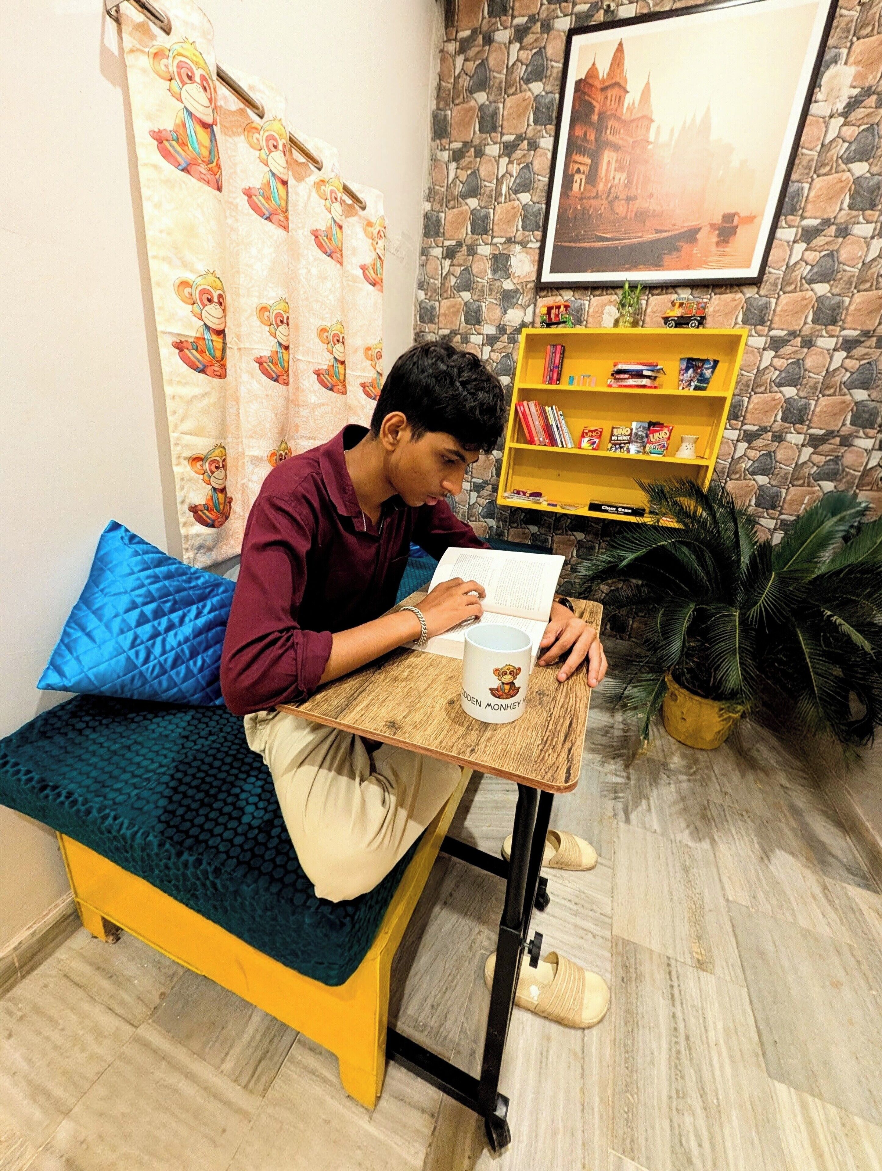 Una persona sentada en un sofá con un libro, una taza y una mesita de noche en una habitación con pared de piedra, estantería y planta en maceta.