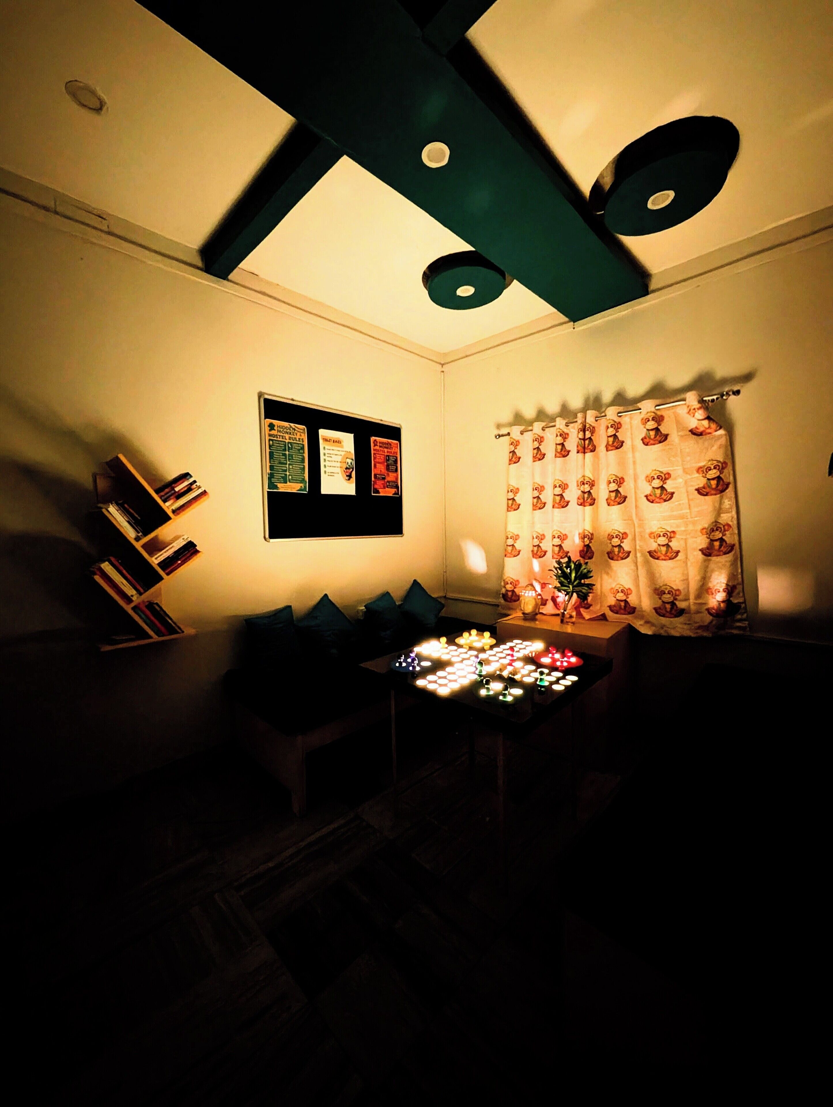 Una habitación tenuemente iluminada con un ventilador de techo, una mesita con velas, una estantería y una cortina con estampado de monos.