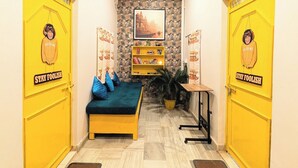 A narrow hallway with yellow doors, a bed with blue pillows, a small table, and a stone wall with a framed picture. - Hidden Monkey Hostel Chet Singh Ghat (Varanasi)