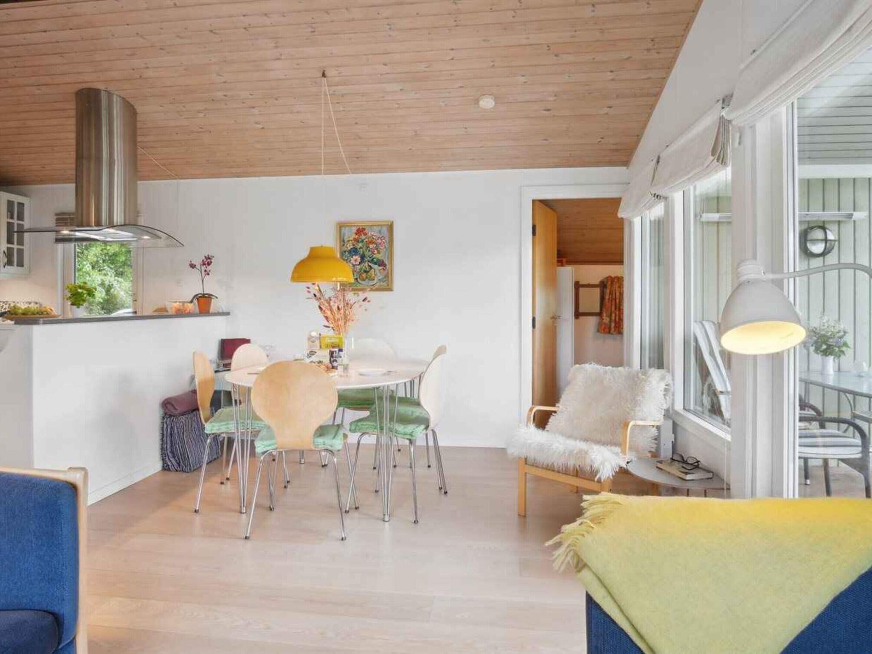 A modern kitchen with wooden ceiling, a dining area with a round table and chairs, and a cozy seating area with a chair and a lamp.