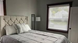 A bedroom with a tufted headboard, a bed with a grey quilt, a bedside table, a floor lamp, and a window with blinds. - Quiet 3 Bedroom home Near downtown (Lansing)