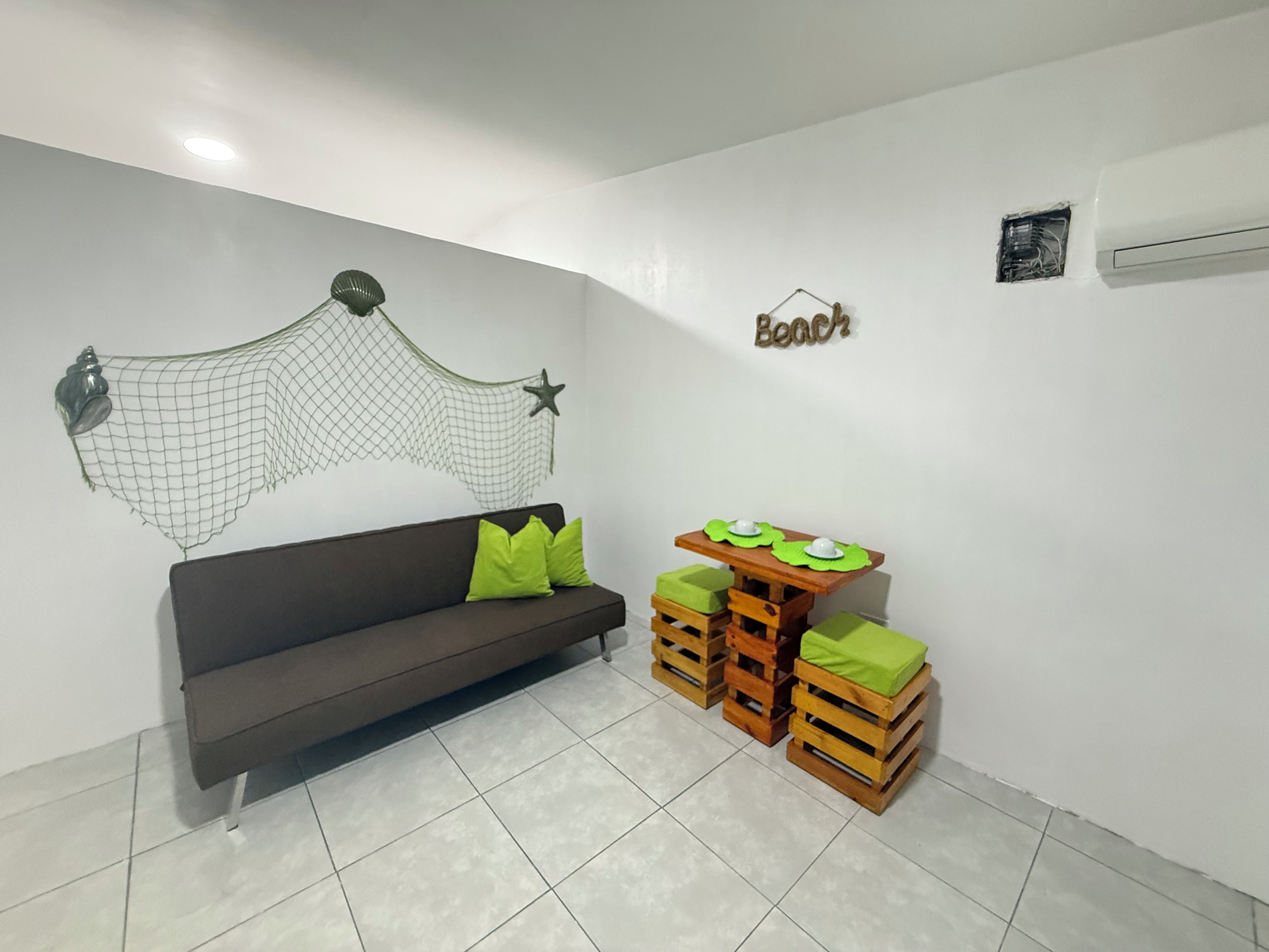 Una habitación con un sofá marrón oscuro, cojines verdes, una pequeña mesa de madera con cojines verdes, una hamaca montada en la pared y un adorno de pared con una estrella y la palabra 'Beach'.