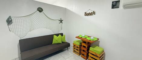 Una habitación con un sofá marrón oscuro, cojines verdes, una pequeña mesa de madera con cojines verdes, una hamaca montada en la pared y un adorno de pared con una estrella y la palabra 'Beach'.