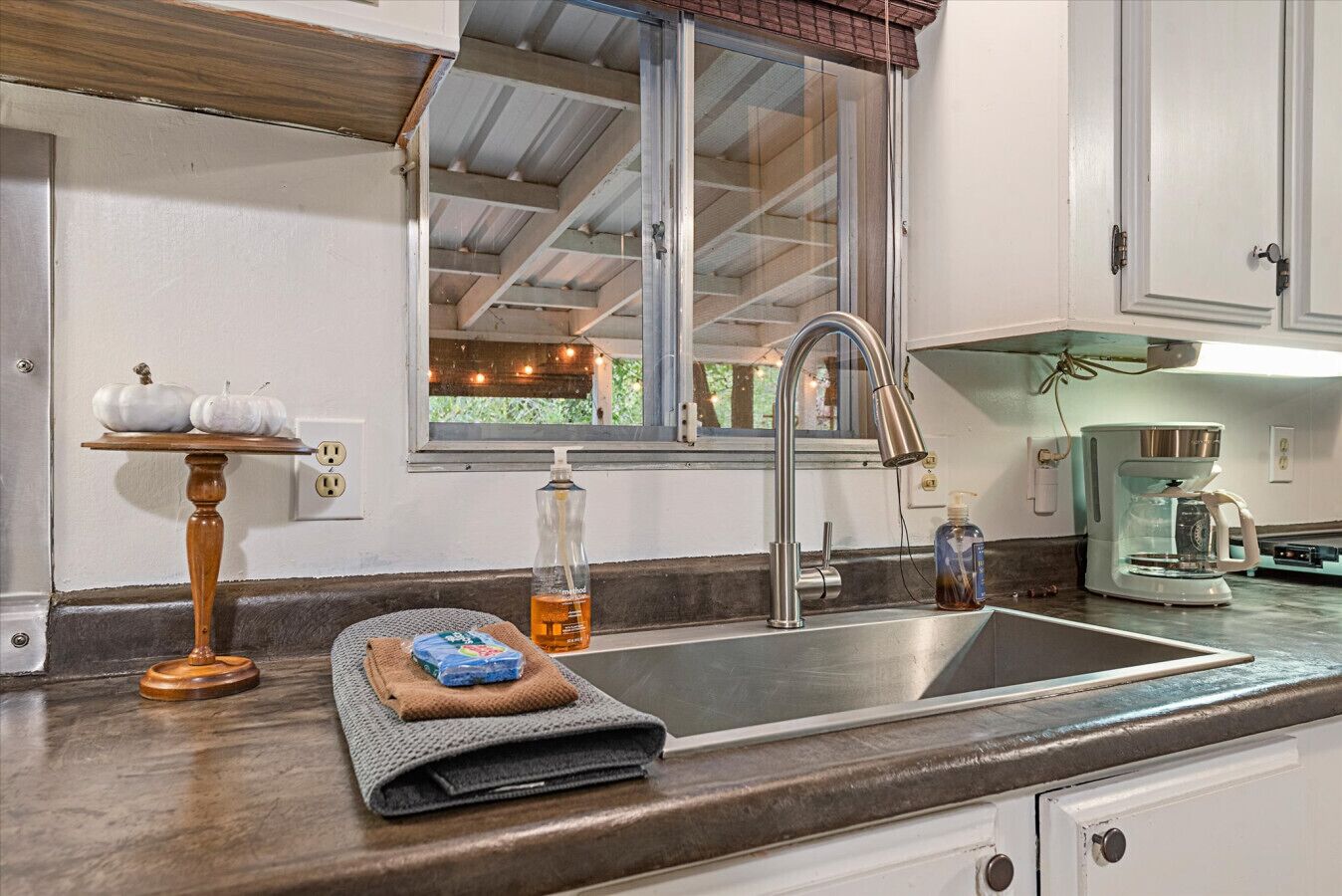 A kitchen with a stainless steel sink, a coffee maker, and a window with a view of greenery.