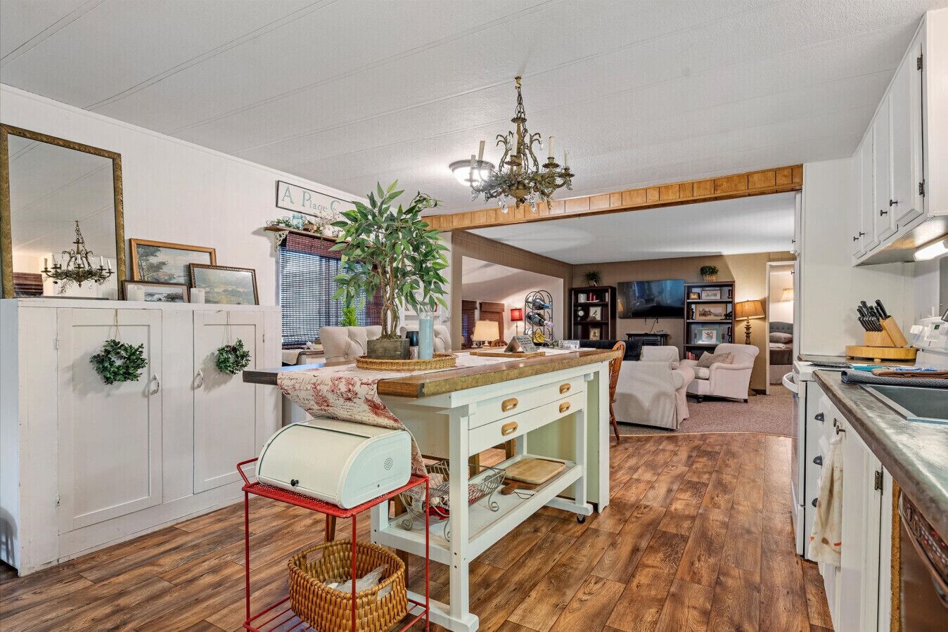 Un vaste espace de vie doté d’un plancher en bois, d’un îlot central avec un évier et d’une table à manger. On y trouve également une cuisine avec des armoires blanches et un lustre.