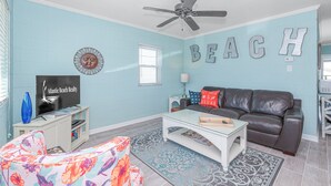 Una sala de estar con una pared azul, un ventilador de techo, un sofá de cuero marrón, una mesa de centro blanca y un sofá floral.