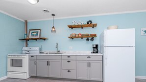 Una cocina con paredes azul claro, un refrigerador blanco, una estufa y un lavamanos. Hay repisas de madera sobre la encimera con vajilla y un marco de fotos.
