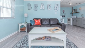 Un salón con una pared azul, una mesa de centro blanca, un sofá gris y una zona de cocina con encimera y armarios blancos.
