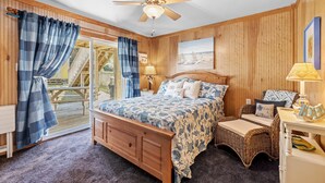 Une chambre avec un ventilateur de plafond en bois, un lit recouvert d’une courtepointe à motifs floraux bleus et blancs, une chaise en osier et une terrasse en bois visible à travers la porte vitrée.
