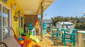 Une terrasse en bois agrémentée de chaises Adirondack colorées et d’une table, un panneau indiquant la direction de la plage, et une décoration en forme de planche de surf.