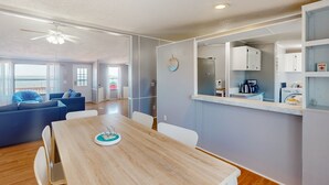Un salon moderne avec une table à manger en bois, des canapés bleus et un coin cuisine doté de meubles blancs et d’appareils électroménagers.