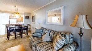 A living room with a striped sofa, a dining table with chairs, a lamp, and a mirror on the wall. - First-Floor Condo in Soundside Complex with WiFi, W/D & Shared Pools/Gym/Tennis (Atlantic Beach)