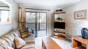 A living room with a sofa, a coffee table, a TV stand, and a sliding glass door leading to a balcony. - First-Floor Condo in Soundside Complex with WiFi, W/D & Shared Pools/Gym/Tennis (Atlantic Beach)