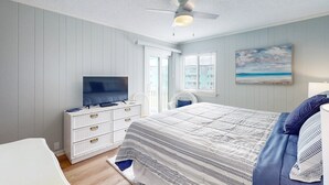 Een slaapkamer met een bed, een televisie op een dressoir, een plafondventilator en een schilderij van een strand.
