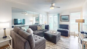 Een moderne woonkamer met een grijze modulaire bank, een grijze fauteuil, een salontafel en een grote spiegel. Er is een plafondventilator, een televisie en een raam met zonwering.