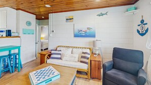 Un salon chaleureux avec un plafond en bois, un canapé, un fauteuil bleu, une table basse sur laquelle est posé un jeu de société, et des œuvres d’art murales sur le thème de la mer.