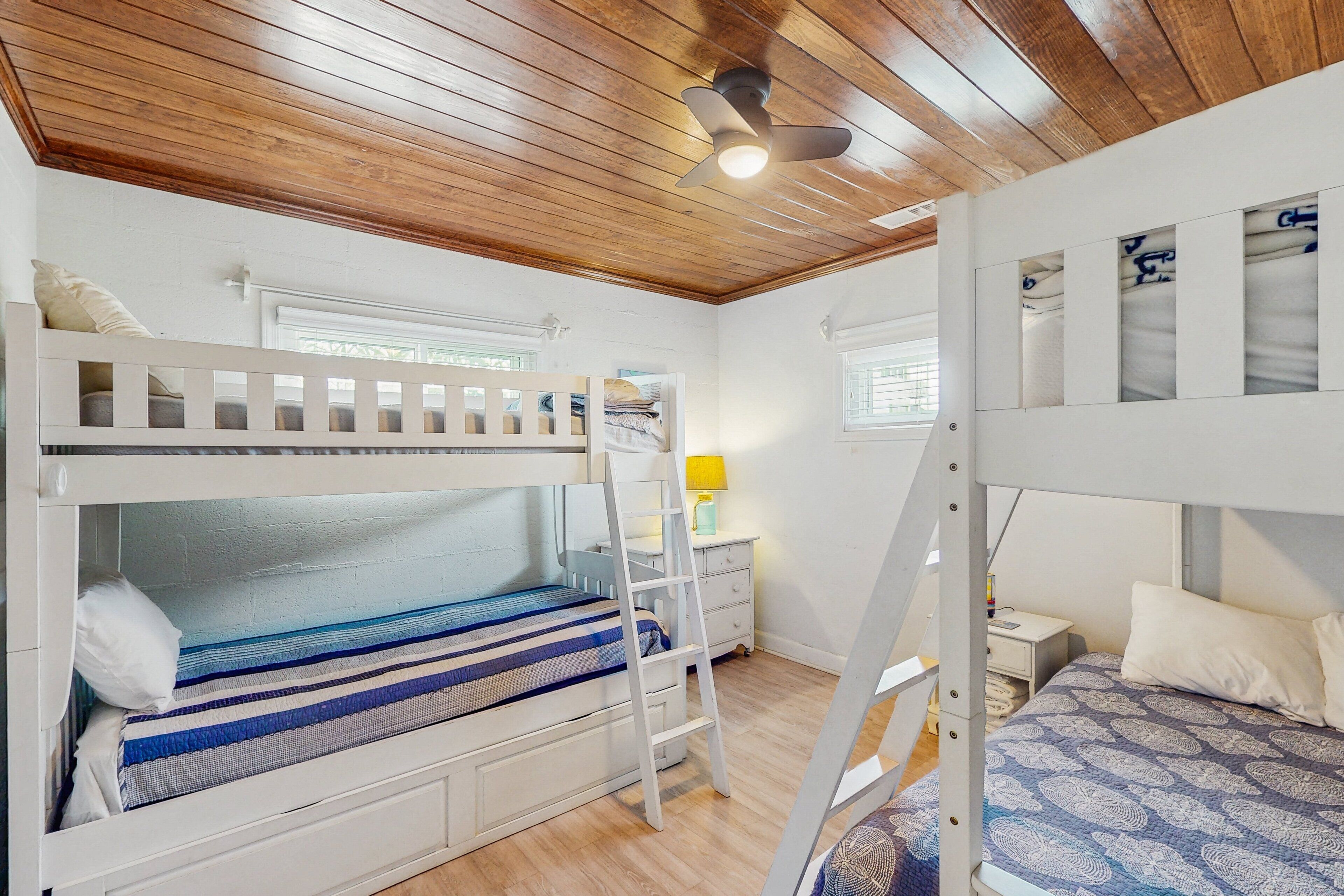 A bunk bed room with a wooden ceiling, a ladder, a bedside table with a lamp, and a window with blinds.