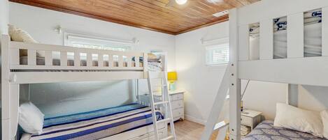 A bunk bed room with a wooden ceiling, a ladder, a bedside table with a lamp, and a window with blinds.