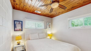 Une chambre avec un plafond en bois, un lit recouvert d’une literie blanche, deux lampes de chevet, une fenêtre avec des stores et un tableau encadré au mur.