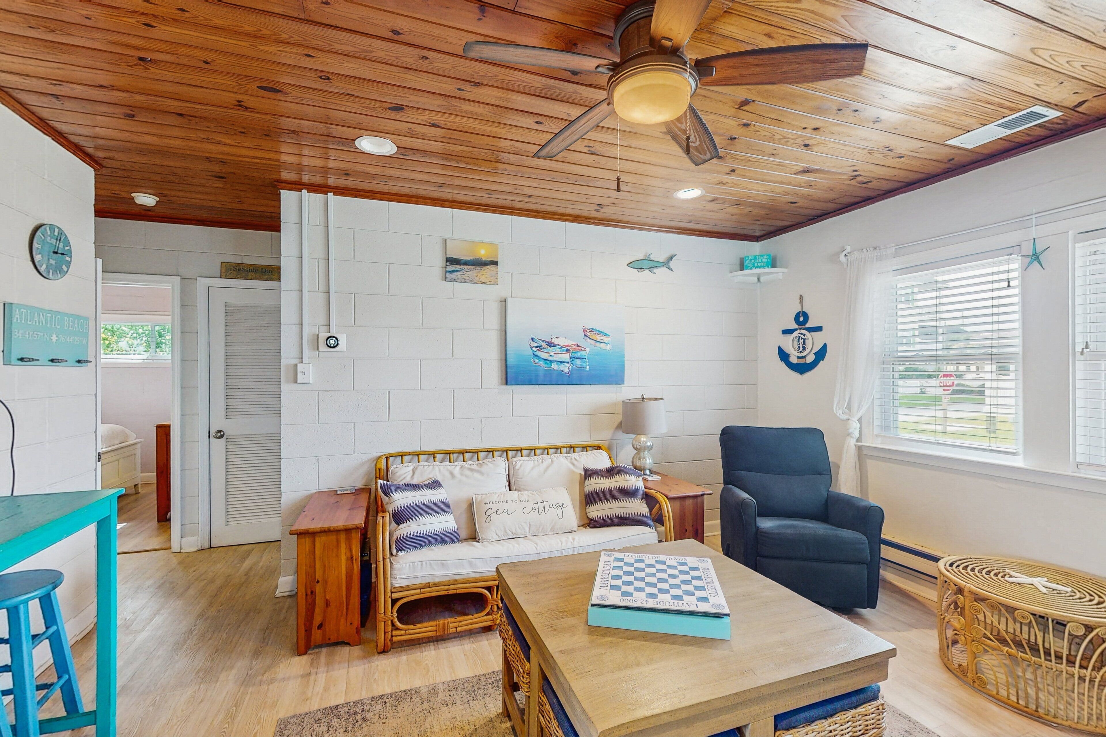 A cozy room with a wooden ceiling, a wicker sofa, a wooden table, and a view of the outdoors through a window with blinds.