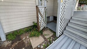 Un espacio al aire libre cubierto con piso de madera, un portón de celosía blanca y un escalón de concreto que lleva a una puerta.