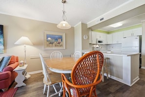 A dining area with a wooden table and chairs, a kitchen with white cabinets and appliances, and a living room with a sofa and a lamp.