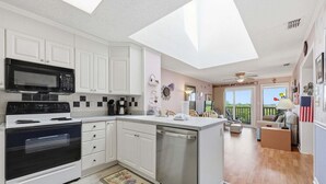 A modern kitchen with white cabinets, a microwave, stove, and dishwasher. There is a balcony with outdoor seating and a view of the sky. - Well-Appointed Condo with WiFi, Washer/Dryer & Shared Pools, Tennis (Atlantic Beach)