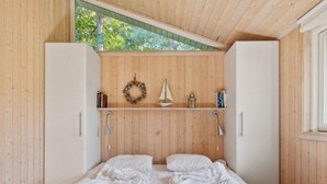 Un dormitorio con techo inclinado de madera, cama con ropa de cama blanca, estante con libros y una guirnalda, además de una pequeña decoración con forma de velero.