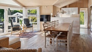 Amplia sala de estar con techo de madera, una mesa de comedor con sillas, televisor y una cocina con armarios y lámpara colgante.