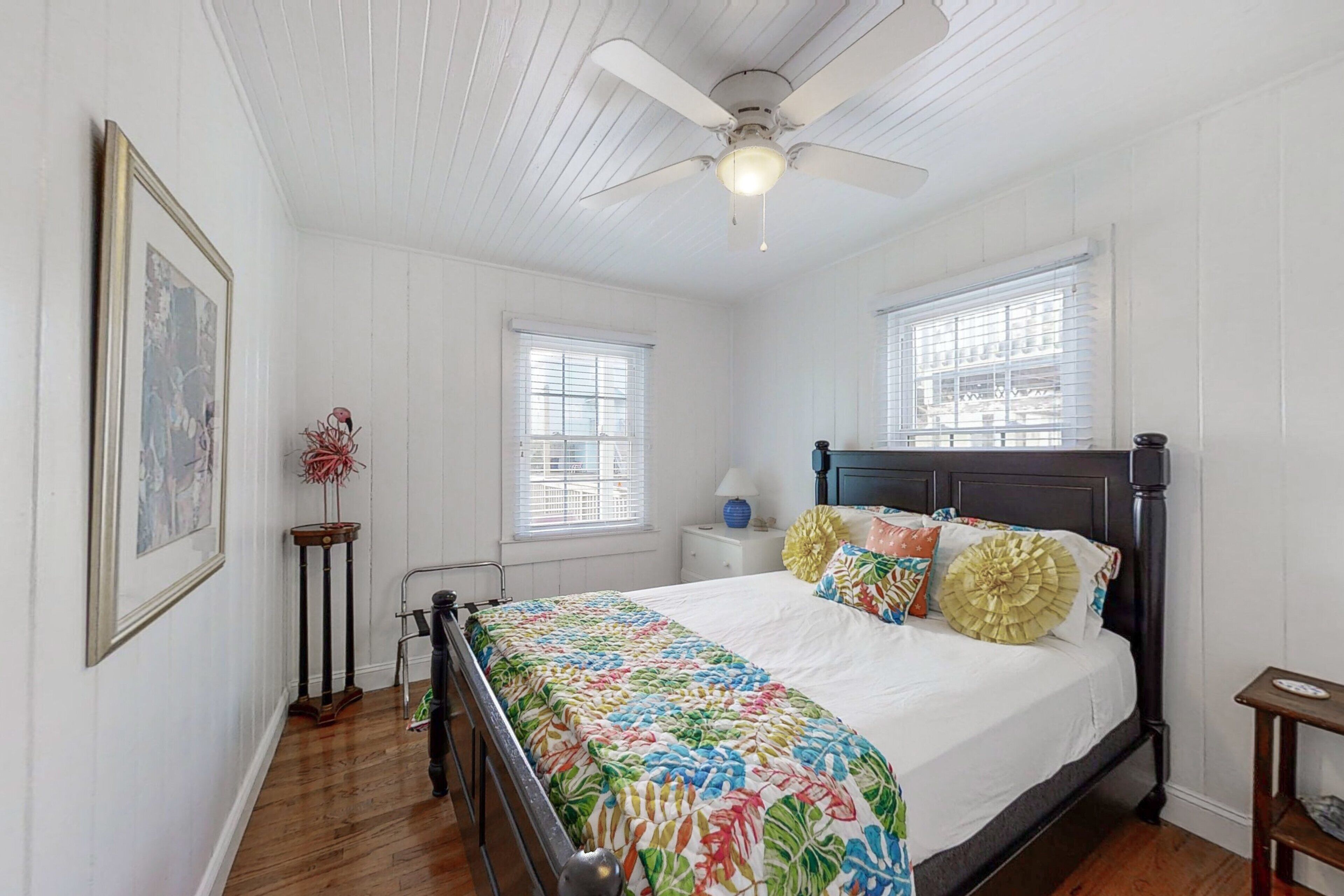 A bedroom with a wooden floor, a bed with a colorful quilt, a ceiling fan, and two windows with blinds.