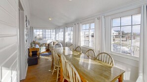 A spacious dining area with a long wooden table and white chairs, large windows with a view of the city, and a small seating area with a table and chairs. - Bullard Cottage: In the heart of Atlantic Beach w/ocean views (Atlantic Beach)