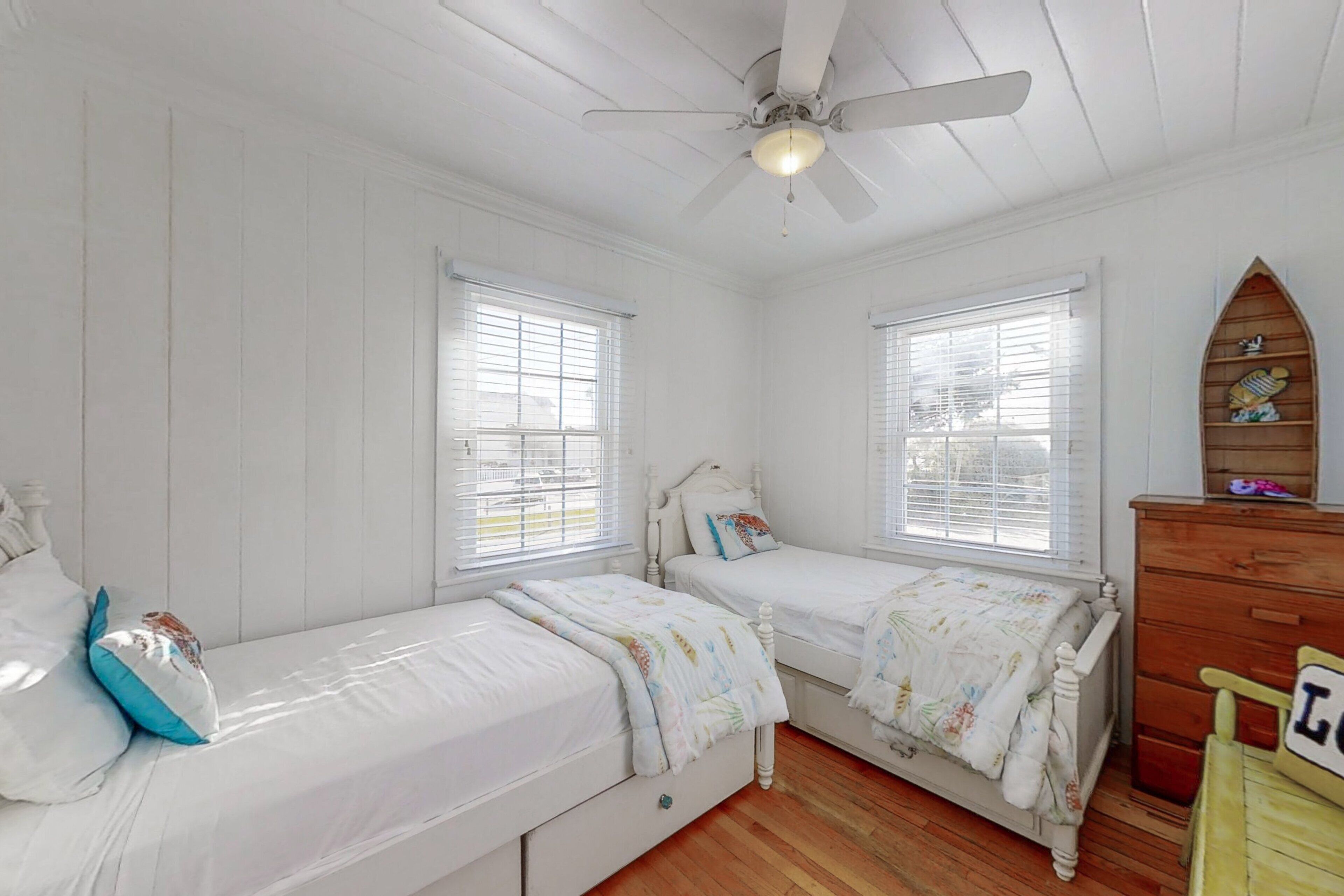 Une chambre avec deux lits simples, une commode en bois et un ventilateur de plafond.