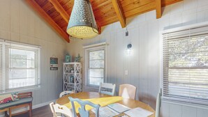 A dining area with a wooden table, white chairs, and a hanging light fixture. - 3rd-row beach cottage with ocean views, beach access, deck, & grill (Emerald Isle)