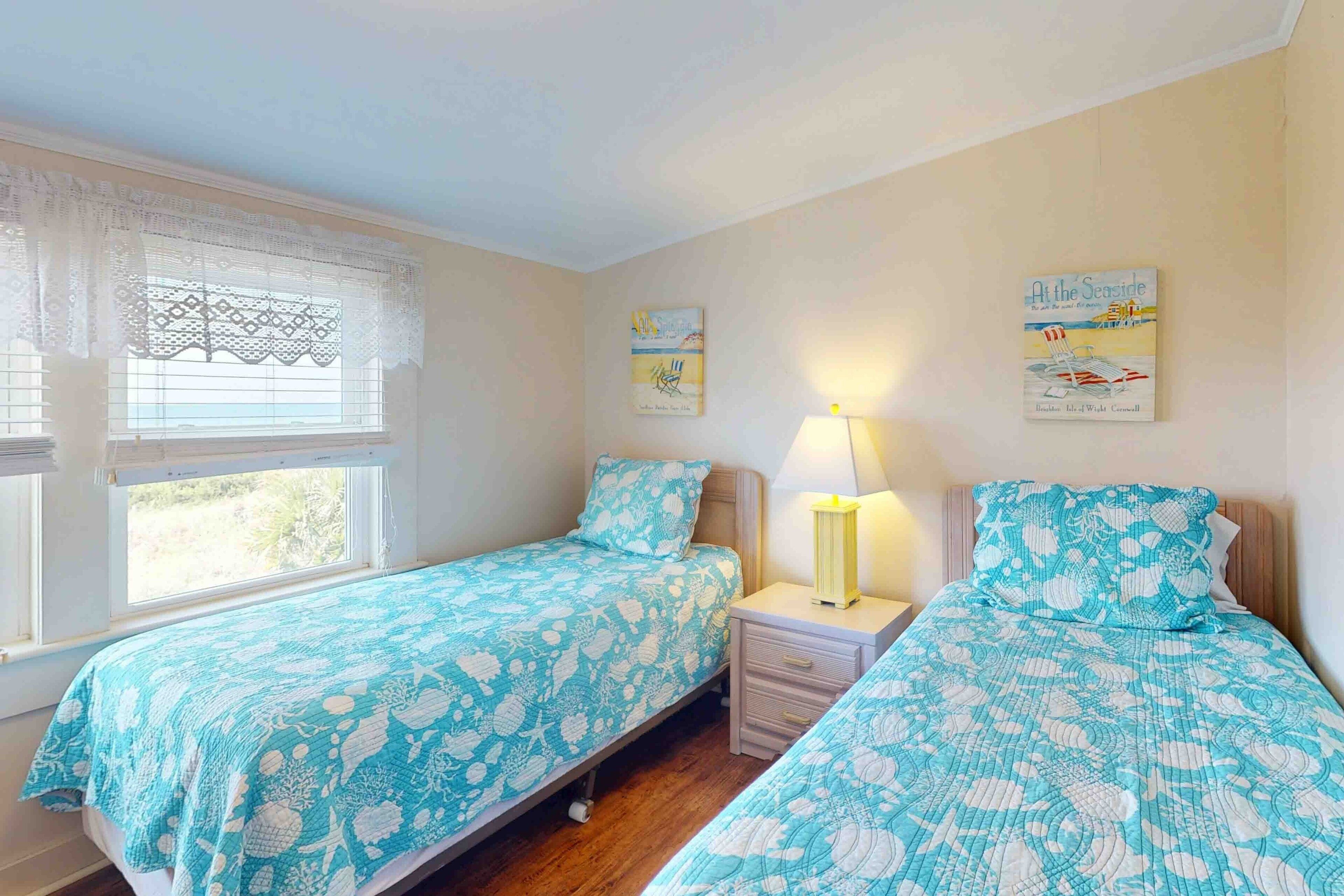 Quarto com duas camas de solteiro, mesa de cabeceira com abajur e janela com cortinas de renda.
