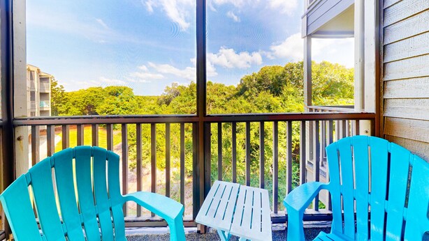 A covered porch with two turquoise chairs and a small table, overlooking a green landscape with trees and a building in the background. - Bright Condo with WiFi, W/D, Screened Porch & Community Pool/Tennis/Day Dock (Atlantic Beach)