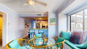 A dining area with a glass-top table surrounded by wicker chairs, a sofa with patterned cushions, and artwork on the walls. - Bright Condo with WiFi, W/D, Screened Porch & Community Pool/Tennis/Day Dock (Atlantic Beach)