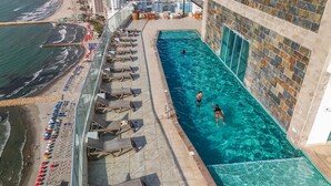 A rooftop pool with clear water, adjacent to a beach with umbrellas and lounge chairs.